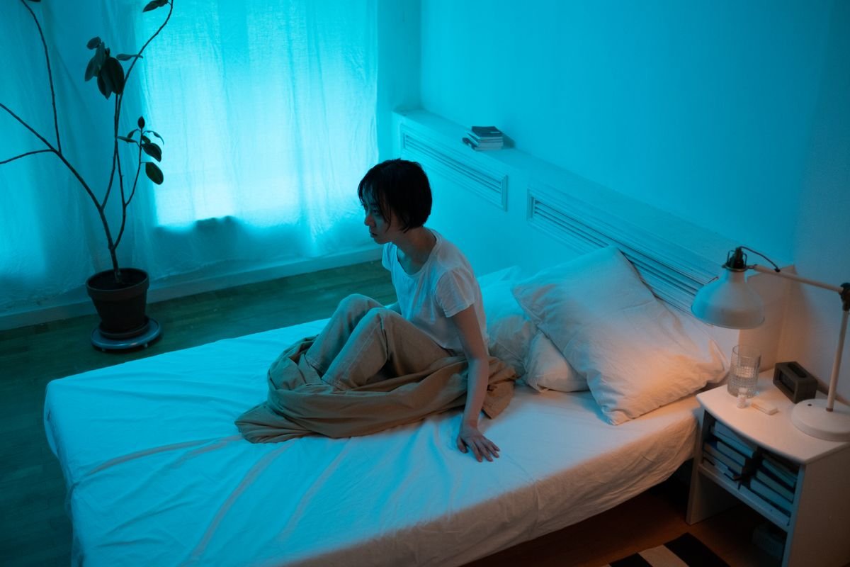 Mulher sentada na cama em quarto com pouca luz, em momento de reflexão