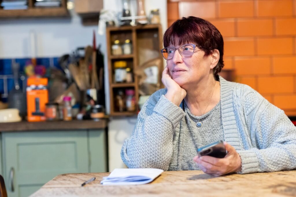 Mulher madura pensativa a mesa da cozinha segurando o celular